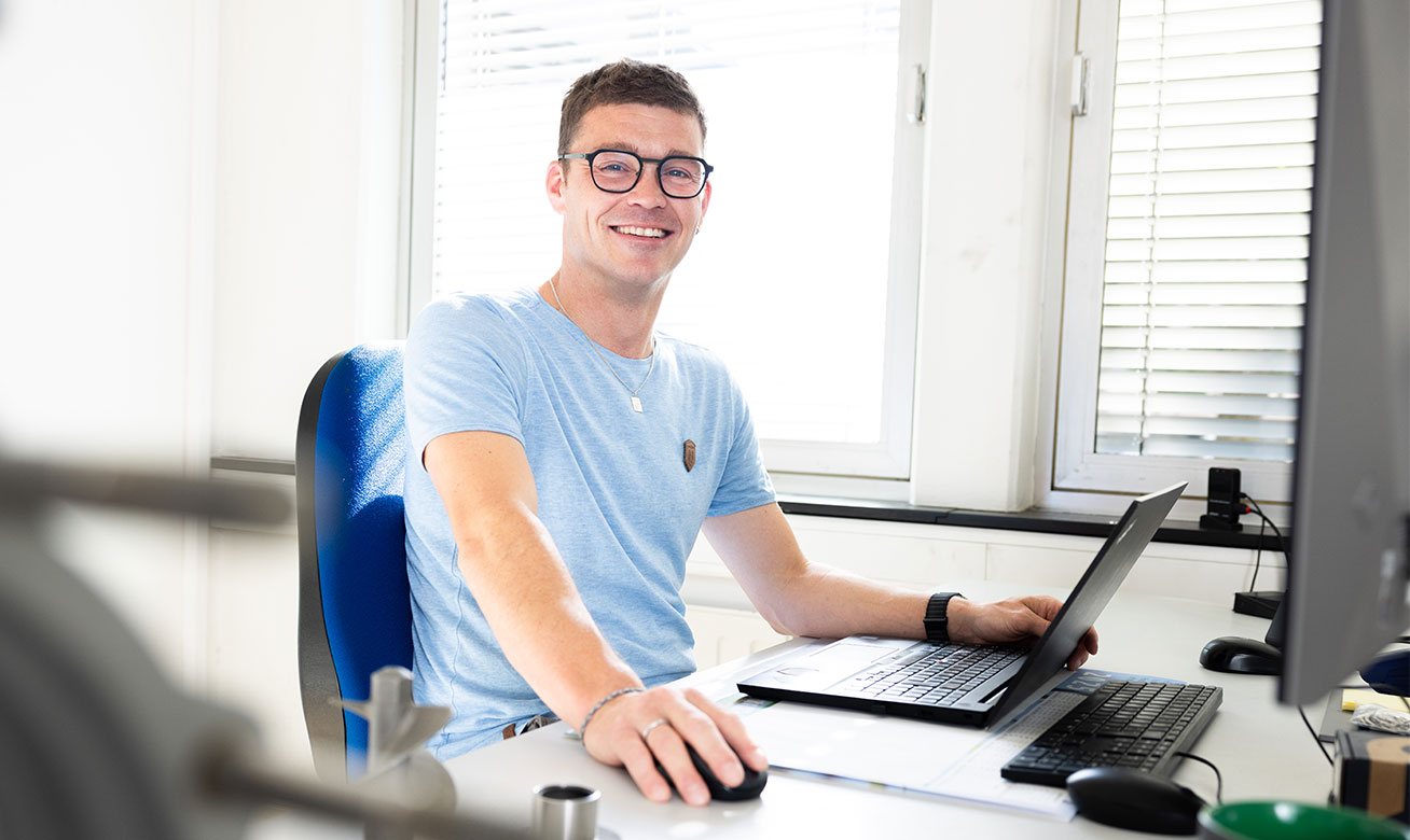 Lächelnder Mann mit Brille sitzt am Schreibtisch vor Laptop und Computer im Büro.
