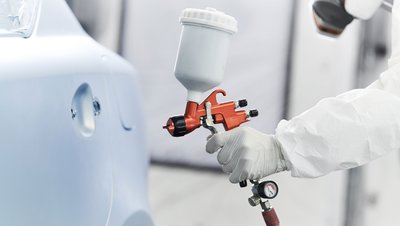 Technician in white protective suit using an orange HVLP spray gun to paint a car body panel.