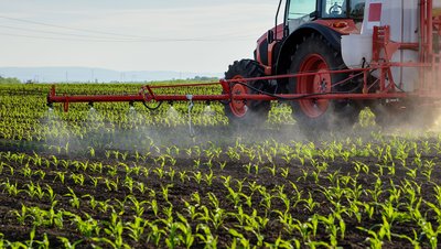 Red tractor with boom sprayer applying liquid fertilizer or pesticide to rows of young corn seedlings in a field.