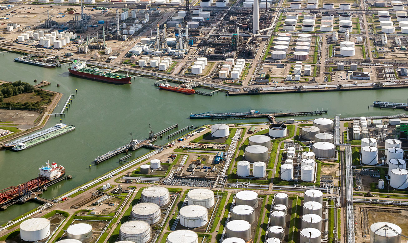 <h2>Safe conveyance of volatile and flammable media</h2> Aerial view of a large petrochemical port with oil storage tanks, refineries, and tanker ships docked along industrial waterways.