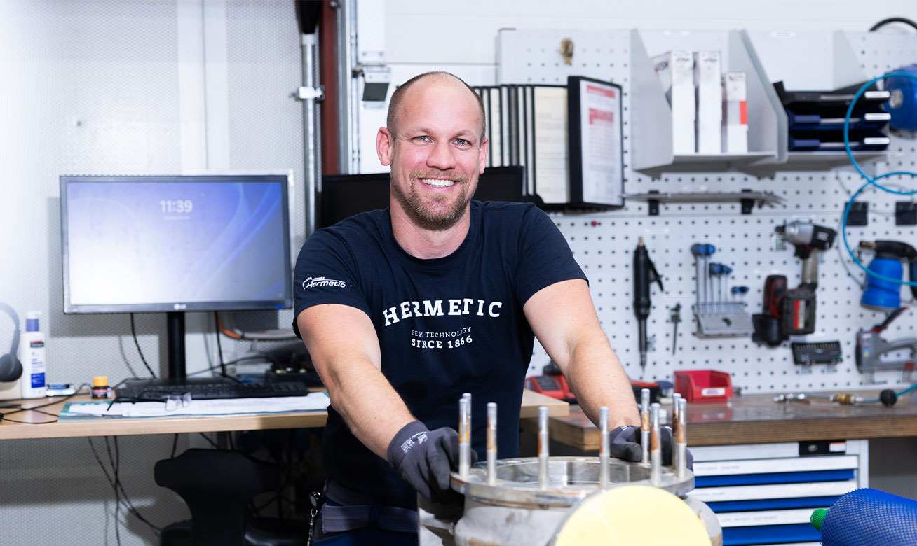 Smiling technician in HERMETIC uniform working on mechanical equipment in a well-organized workshop with tools on pegboard.