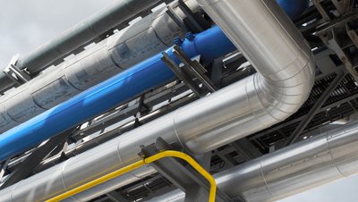 <h3>Pumping of heat transfer oil for heating reactors and heat exchangers</h3> Industrial pipeline system with insulated silver, blue, and yellow pipes on a steel support structure against cloudy sky.