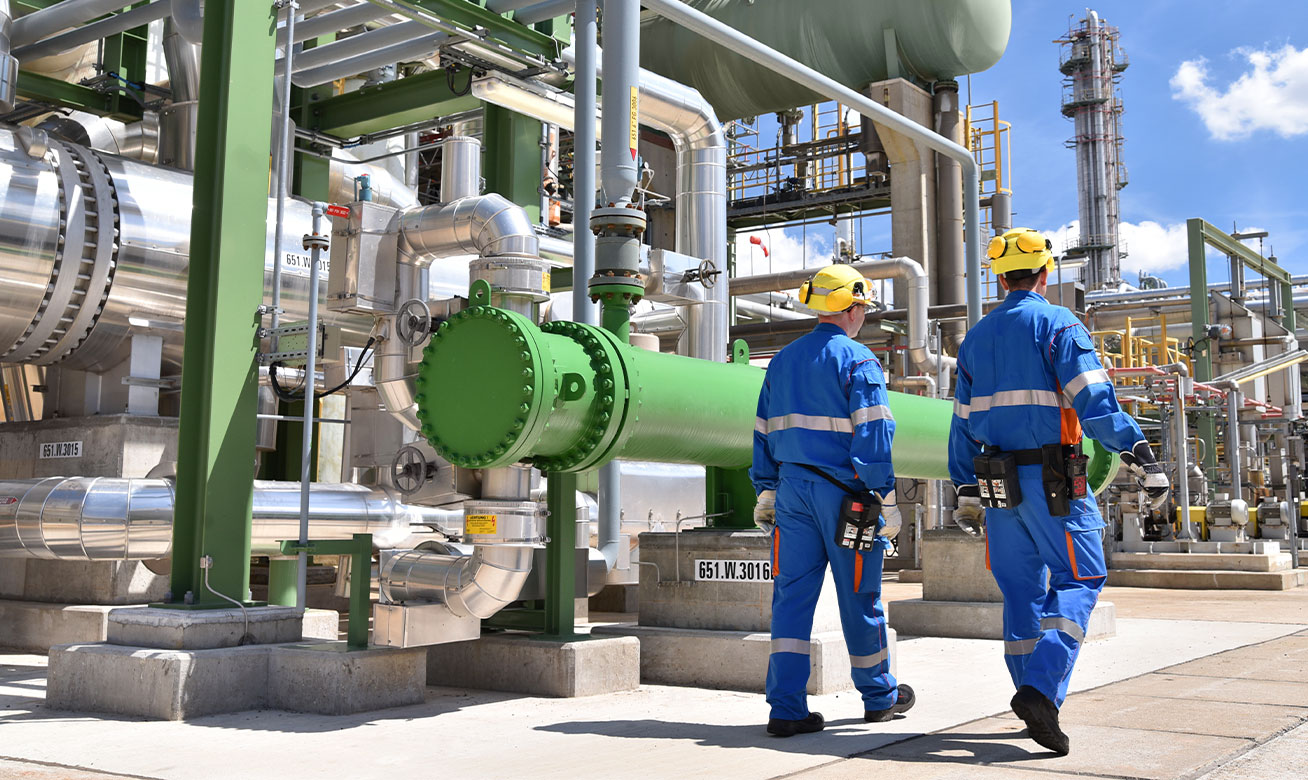 Two workers in blue coveralls and yellow hard hats inspecting pipelines and equipment at a chemical plant.