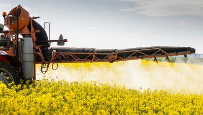 Agricultural sprayer applying mineral acid-based pesticides over a blooming yellow rapeseed field.