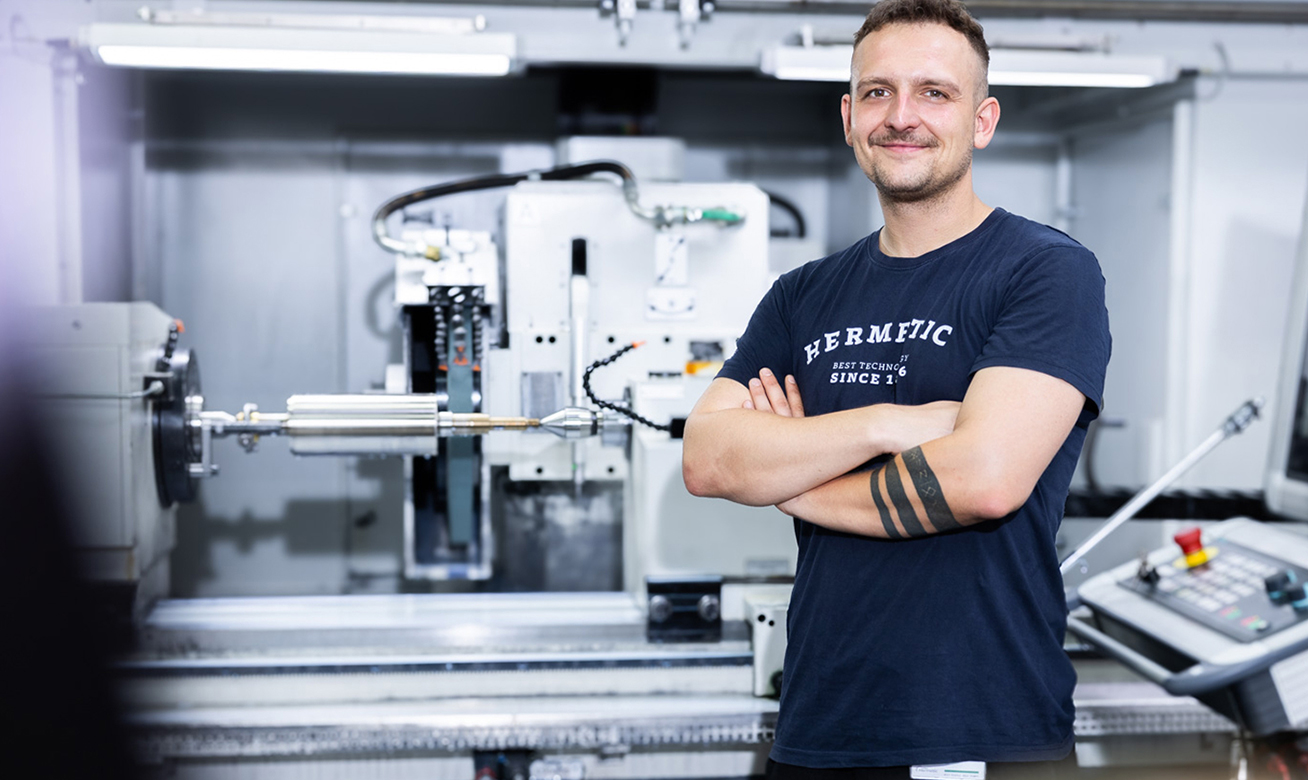 Smiling male worker with crossed arms wearing a HERMETIC t-shirt standing in front of CNC machinery in a factory.