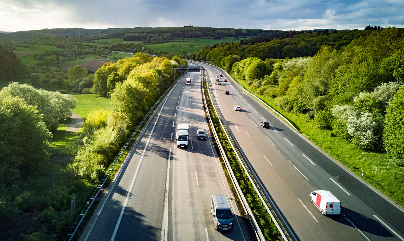 Aerial view of a dual carriageway highway surrounded by lush green forests and rolling hills, with cars and trucks in traffic.