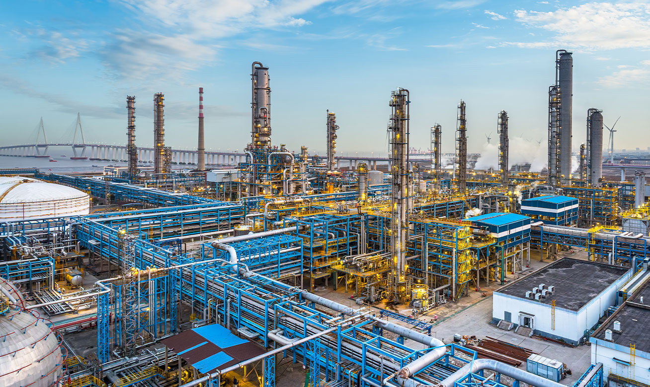 Aerial view of a large petrochemical refinery with blue pipelines, distillation towers, and golden lights near a waterway.